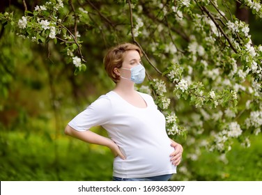 Portrait of a beautiful pregnant young woman wearing disposable medical face mask in the spring park during coronavirus outbreak. Safety in a public place while epidemic of covid-19.