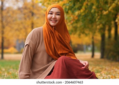 Portrait of beautiful pretty Asian Muslim young woman , Islamic girl is wearing on head hijab scarf sitting on grass, colourful leaves in golden autumn, fall park. Traditional Muslim Arabian clothes