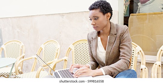 Portrait Of Beautiful Professional African American Office Female In Coffee Shop Using A Laptop Computer, Working In City Outdoors. Black Business Woman With Technology And Paperwork, Exterior.