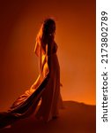 portrait of beautiful red-haired woman wearing long flowing fantasy toga gown with golden halo crown jewellery,  creative hand gestures on a dark moody background with glowing orange lighting.