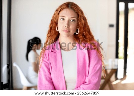 Portrait of beautiful redhead business woman or office worker. A young and dynamic professional stands in a white-walled room. She exudes seriousness, wearing a pink blazer.