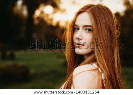 Portrait of a beautiful redhead woman with green eyes and freckles looking at camera against sunset outside over the shoulder.