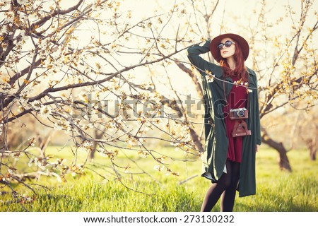 Portrait of a beautiful redhead women in cloak and hat with camera in blossom apple tree garden in spring time on sunset.