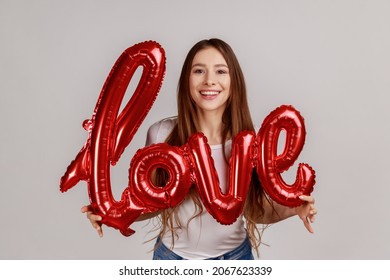 Portrait Of Beautiful Satisfied Romantic Woman Holding Foil Balloon Letters, Looking At Camera, Expressing Love, Wearing White T-shirt. Indoor Studio Shot Isolated On Gray Background.