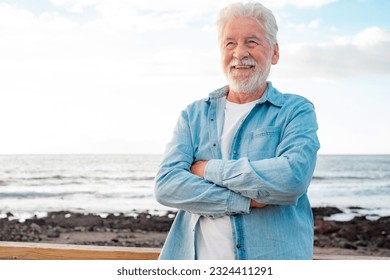 Portrait of beautiful senior bearded man in denim shirt standing outdoors at sea in sunset light looking away. Smiling old man relaxed enjoying retirement - Powered by Shutterstock