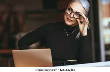 Portrait of beautiful senior businesswoman sitting at cafe with laptop looking away and thinking. Mature woman at coffee shop. - Powered by Shutterstock