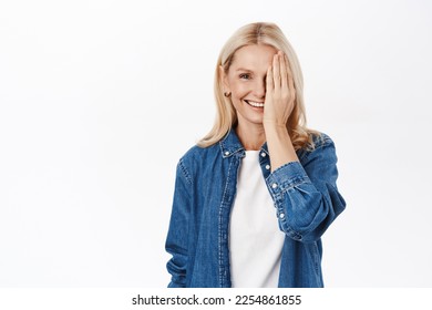 Portrait of beautiful senior woman with blond hair, cover half of face, one side, smiling and looking at camera happy, standing over white background - Shutterstock ID 2254861855