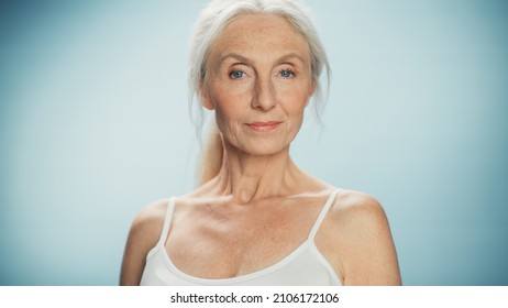 Portrait Of Beautiful Senior Woman Looking At Camera And Smiling Wonderfully. Elderly Lady With Natural Lush Grey Hair, Blue Eyes, Wearing Top. Beauty And Dignity Of Old Age