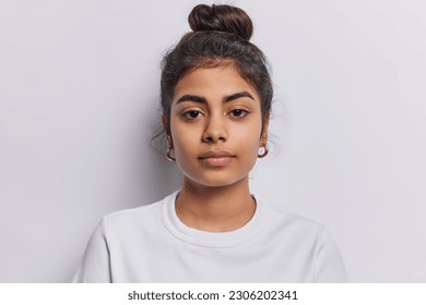 Portrait of beautiful serious brunette woman focused at camera has dark hair combed in bun dressed in casual t shirt isolated over white background. Confident Idian female model with calm expression