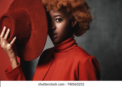 Portrait Of Beautiful Sexy Black Female Model Wearing Red Sweater And Jacket, Holding Red Hat In Her Hand In Studio With Grey Background