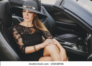 Portrait Of Beautiful Sexy Fashion Woman Model In Summer Hat And In Black Dress And Luxury Accessories With Bright Makeup Sitting In Luxury Car. Young Woman Driving On Road Trip On Sunny Summer Day.