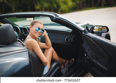 Portrait of beautiful sexy fashion woman model in summer hat and in black dress and luxury accessories with bright makeup sitting in luxury car. Young woman driving on road trip on sunny summer day.