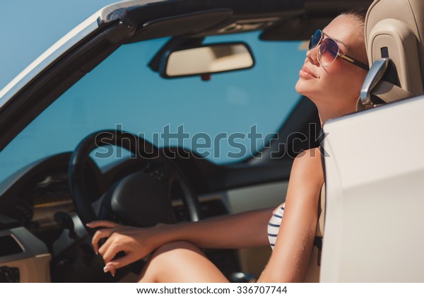 Portrait of beautiful sexy fashion woman model in\
sunglasses sitting in luxury white car with sea and sky background.\
Young woman driving on road trip on sunny summer day. Sea and sky.\
White cabrio.