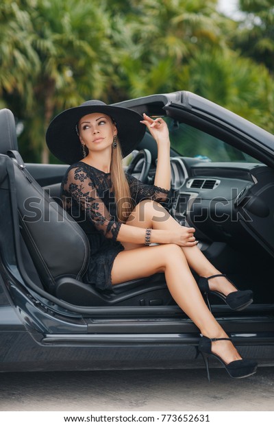 Portrait of beautiful sexy fashion woman model in\
summer hat and in black dress and luxury accessories with bright\
makeup sitting in luxury car. Young woman driving on road trip on\
sunny summer day.