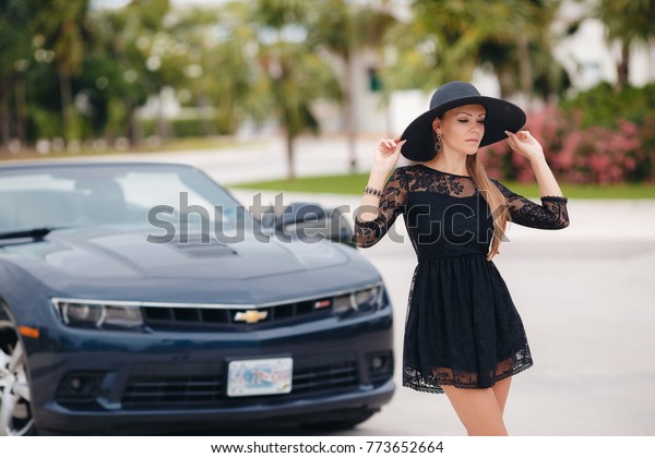 Portrait of beautiful sexy fashion woman model in\
summer hat and in black dress and luxury accessories with bright\
makeup sitting in luxury car. Young woman driving on road trip on\
sunny summer day.