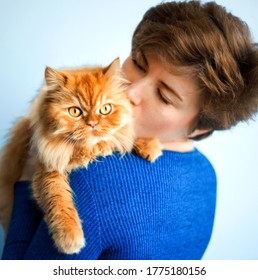 Portrait Of Beautiful Short Haired 30 Years Old Woman In Blue Dress Kissing Her Ginger Red Furry Persian Cat. Domestic Animal, Pet And Host Together. People Love Kitten. Cat Day.