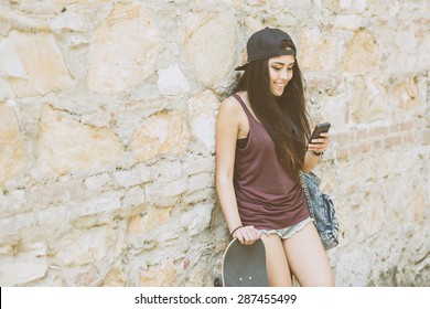 Portrait Of A Beautiful Skater Girl Looking At Smart Phone Against Stone Wall. She Is Half Caucasian And Half Filipina, She Wears Short Jeans, A Purple Tank Top And A Black Cap.