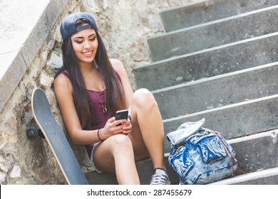 Portrait Of A Beautiful Skater Girl Looking At Smart Phone At Park. She Is Half Caucasian And Half Filipina, She Wears Short Jeans, A Purple Tank Top And A Black Cap.