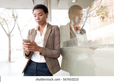 Portrait Of Beautiful Slick African American Business Woman With Shiny Office Building City Reflections, Thoughtful, Holding Smart Phone Outdoors. Professional Black Woman Using Technology, Lifestyle.