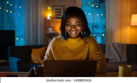 Portrait Of Beautiful Smiling Black Girl Working On A Laptop While Sitting At Her Desk At Home. In The Evening Creative Woman Works On A Computer In Her Cozy Living Room.