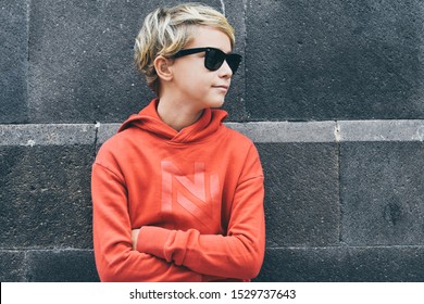 Portrait Of A Beautiful Smiling Boy With Sunglasses Leaning On A Black Wall. Trendy Blond Teen With Red Sweatshirt Looking Away Arms Crossed. Serious Young Male. Youth, Relax Fashion People Concept