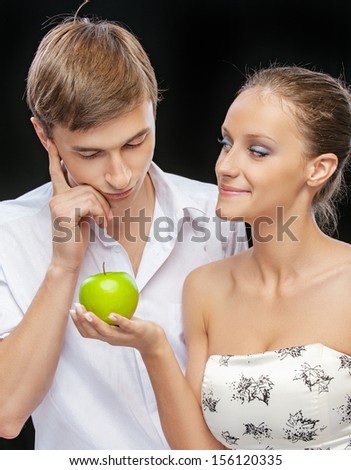 portrait of beautiful smiling brunette girl giving apple to confused happy guy on black