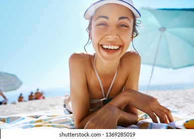 portrait of beautiful smiling happy woman tanning in bikini and white cap on sandy beach at summer. Model is relaxing lying down on white sand tropical getaway. Summer vacation concept.