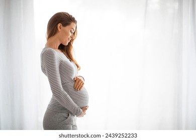 Portrait Of Beautiful Smiling Pregnant Woman Tenderly Embracing Belly While Standing Against Window At Home, Happy Expectant Mother Enjoying Her Pregnancy Time, Side View With Copy Space - Powered by Shutterstock