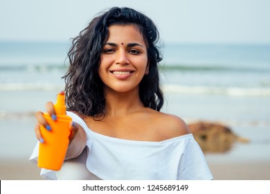 portrait of a beautiful and smiling snow-white smile indian woman black curly hair and dark skin in a white t-shirt holding bottle of sunscreen spray on beach.girl enjoying spf body paradise vacation