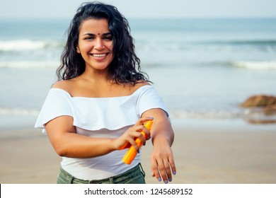 Portrait Of A Beautiful And Smiling Snow-white Smile Indian Woman Black Curly Hair And Dark Skin In A White T-shirt Holding Bottle Of Sunscreen Spray On Beach.girl Enjoying Spf Body Paradise Vacation