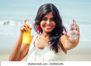 Portrait Of A Beautiful And Smiling Snow-white Smile Indian Woman Black Curly Hair And Dark Skin In A White T-shirt Holding Bottle Of Sunscreen Spray On Beach.girl Enjoying Spf Body Paradise Vacation