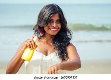 Portrait Of A Beautiful And Smiling Snow-white Smile Indian Woman Black Curly Hair And Dark Skin In A White T-shirt Holding Bottle Of Sunscreen Spray On Beach.girl Enjoying Spf Body Paradise Vacation