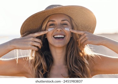 Portrait of beautiful smiling woman applying sunscreen on her face while looking at camera at the beach. - Powered by Shutterstock