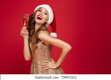 Portrait Of A Beautiful Smiling Woman In Christmas Hat And Dress Holding Champagne Glass While Standing And Looking Up Isolated Over Red Background