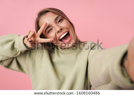 Portrait of beautiful smiling woman gesturing peace sign and taking selfie isolated over pink background