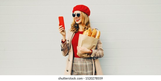 Portrait Beautiful Smiling Woman Holding Phone, Grocery Shopping Paper Bag With Long White Bread Wearing Red Beret Over Blank Gray Wall Background