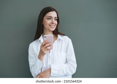 Portrait Of Beautiful Smiling Woman Student With Dark Long Hair In White Shirt Using Mobile Phone In The Hand On Grey Background Isolated, Generation Z