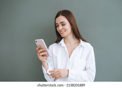 Portrait Of Beautiful Smiling Woman Student With Dark Long Hair In White Shirt Using Mobile Phone In Hand On Grey Background Isolated, Generation Z