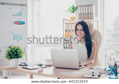 Portrait of beautiful smiling young brunette businesswoman sitting at bright modern work station and typing on laptop