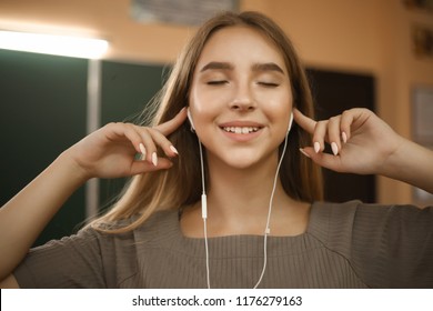 Portrait Of A Beautiful Smiling Young Student Schoolgirl Lady With Long Hair Standing In Empty Classroom Listening Music With Earphones.