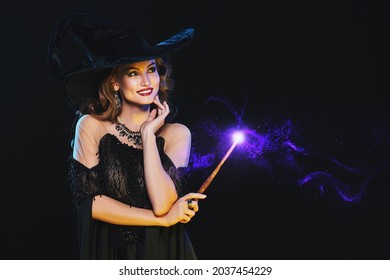 Portrait of a beautiful smiling young witch in a hat and with a magic wand on a black background with magic lights. Halloween celebration, party. Copy space.