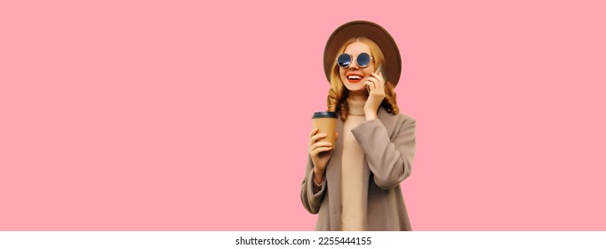 Portrait of beautiful smiling young woman calling on smartphone with cup of coffee wearing brown coat, round hat looking away on pink background, blank copy space for advertising text - Shutterstock ID 2255444155