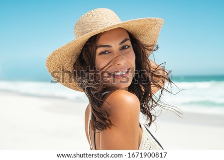 Portrait of beautiful smiling young woman wearing straw hat at beach with sea in background. Beauty fashion girl looking at camera at seaside. Carefree tanned woman walking on sand and laughing.