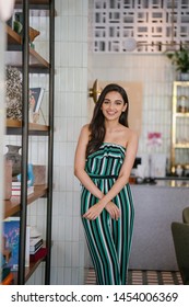 Portrait Of A Beautiful, Sophisticated And Attractive Young Indian Asian Woman (high Society) In An Elegant Green Outfit Smiling As She Stands Next To A Bookshelf In Her Well-appointed Home.