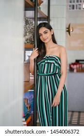 Portrait Of A Beautiful, Sophisticated And Attractive Young Indian Asian Woman (high Society) In An Elegant Green Outfit Smiling As She Stands Next To A Bookshelf In Her Well-appointed Home.