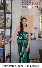 Portrait Of A Beautiful, Sophisticated And Attractive Young Indian Asian Woman (high Society) In An Elegant Green Outfit Smiling As She Stands Next To A Bookshelf In Her Well-appointed Home.