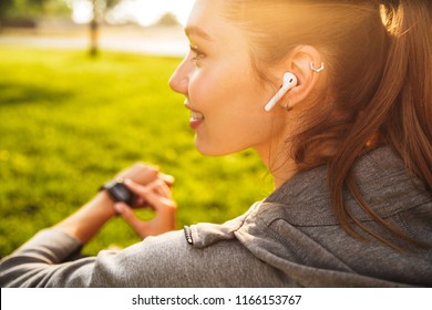 Portrait of beautiful sporty woman 20s in sportswear using smartwatch and wireless earbud while resting in green park