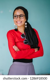 A portrait of a beautiful and stylish Muslim woman in a hijab head scarf and a colorful outfit against in a studio. She is wearing spectacles and smiling happily.  - Shutterstock ID 1286918500