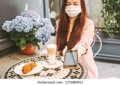 Portrait Beautiful Stylish Woman In Medical Face Mask Use Smart Phone For Contactless Payment In Outdoor French Cafe. Young Girl In Protective Face Mask Drink Coffee, Pay With NFC Technology. COVID-19