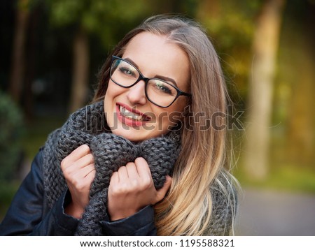 portrait of a beautiful stylish young blonde girl with make-up in black glasses leather jacket and knitted scarf freezes up smiles in the autumn cold park